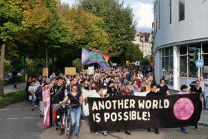 Der Demozug geht gerade auf die Eiserne Brücke. Vorne ist das Frontbanner zu erkenne, auf dem steht " Another World Is Possible" und dahinter befindet sich eine bunte FFF-Fahne und sehr viele Menschen.