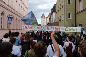 Im Hintergrund ist der Demozug zu erkennen. Im Vordergrund hält eine Person ein Schild, auf dem geschrieben steht "Change the system not the climate".